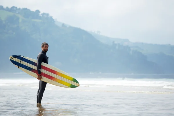 Surfista jovem atraente segurando prancha de surf enquanto está de pé na praia olhando para o oceano para encontrar o local perfeito para ir surfar ondas, surfista profissional com prancha de surf olhando para o oceano, imagem filtrada — Fotografia de Stock