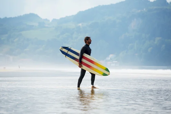 Attraente surfista professionista che tiene la sua tavola da surf a piedi sulla spiaggia, belle passeggiate surfisti portando la sua tavola da surf con oceano sullo sfondo, immagine filtrata — Foto Stock