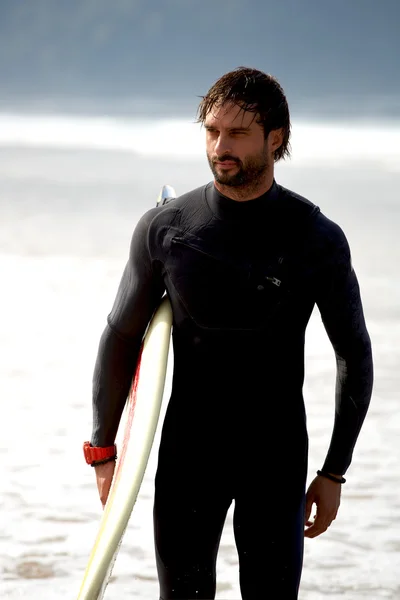 Привлекательный молодой серфер держа доску для серфинга, стоя на пляже глядя на океан, чтобы найти идеальное место для серфинга волны, профессиональный серфер с доской для серфинга глядя на океан, отфильтрованное изображение — стоковое фото