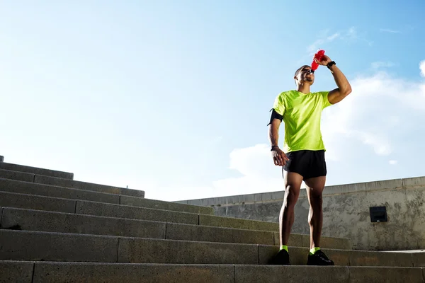 Mörkhyade löpare dricka vatten efter intensiv kväll kör, vackra passar man i ljusa fluorescerande sportkläder, sport fitness koncept — Stockfoto