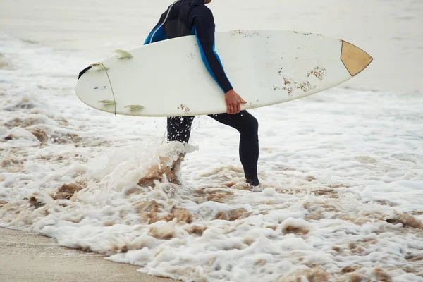 Atractivo surfista profesional sosteniendo su tabla de surf caminando por la playa, apuesto surfista camina llevando su tabla de surf con el océano en el fondo, imagen filtrada — Foto de Stock