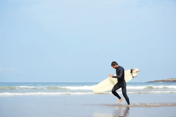 Fiatal, vonzó szörfös fut szörfdeszkával a part mentén, hideg évszak szörfözik az óceán partján, profi szörfös férfi búvárruhában készen a szörfözésre fut a hullámok felé. — Stock Fotó