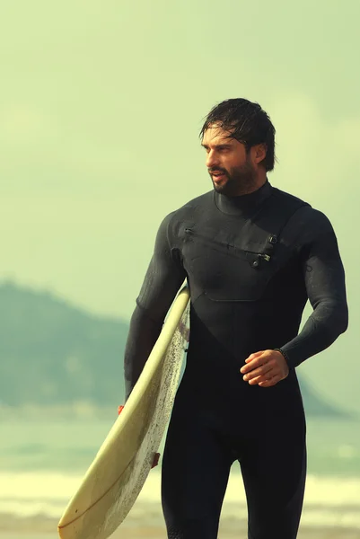 Attraktiver professioneller Surfer, der sein Surfbrett am Strand hält, gutaussehender Surfer, der sein Surfbrett mit Ozean auf dem Hintergrund trägt, gefiltertes Bild — Stockfoto