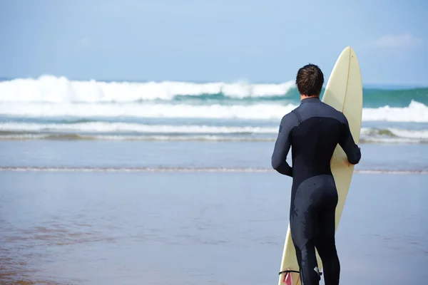 Atractivo joven surfista sosteniendo tabla de surf mientras está de pie en la playa mirando al océano para encontrar el lugar perfecto para ir olas de surf, surfista profesional con tabla de surf mirando al océano, imagen filtrada —  Fotos de Stock