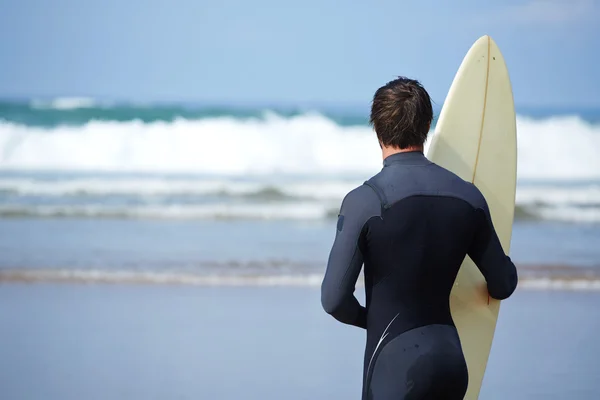 Atractivo joven surfista sosteniendo tabla de surf mientras está de pie en la playa mirando al océano para encontrar el lugar perfecto para ir olas de surf, surfista profesional con tabla de surf mirando al océano, imagen filtrada — Foto de Stock