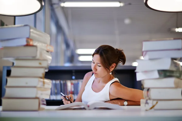 Asiatique étudiante assis à la table avec une énorme pile de livre d'étude et avec téléphone portable à la main — Photo