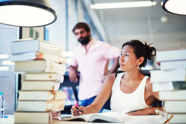 Jonge Aziatische vrouw op Examenvoorbereiding met klasgenoot permanent in de buurt van boekenplank in studie hall — Stockfoto