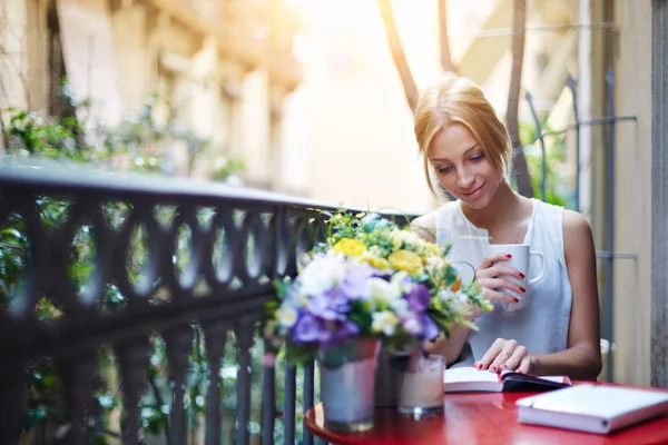 Gadis muda yang menarik membaca buku sambil minum kopi di hari yang cerah duduk di balkon — Stok Foto