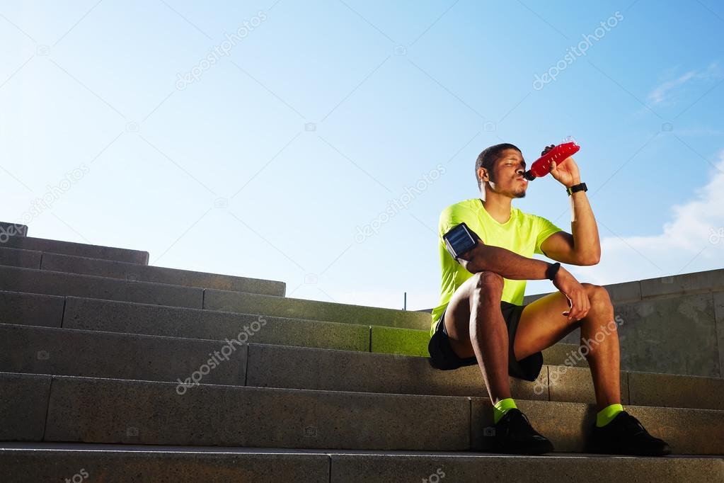 Handsome athletic runner seated on the steps drink water, beautiful fit man in bright fluorescent sportswear, sports fitness concept