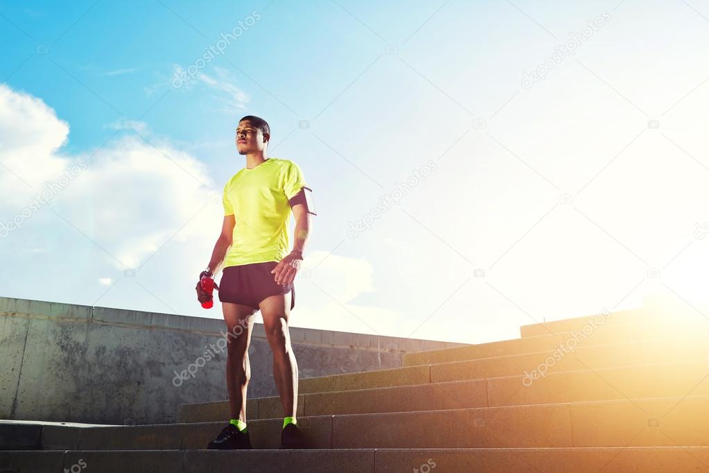 Young dark skinned jogger with muscular strong standing against soft sunset light outdoors, beautiful fit man in bright fluorescent sportswear, sports fitness concept