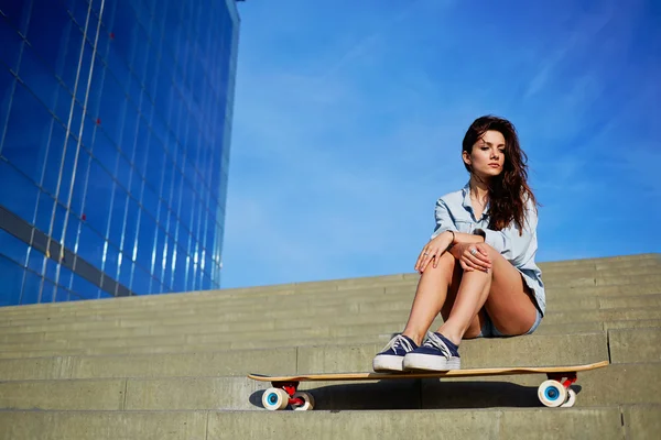 Beautiful sexy girl sits on the steps with big skyscraper on background, young hipster girl posing at beautiful sunset light looking away, attractive woman enjoying sunny evening of summer day — Stock Photo, Image