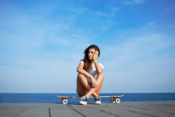 Menina sexy bonita senta-se no longboard com incrível horizonte de mar no fundo, jovem garota hipster posando na bela luz do sol sentado em seu skate, mulher atraente desfrutando de noite ensolarada — Fotografia de Stock