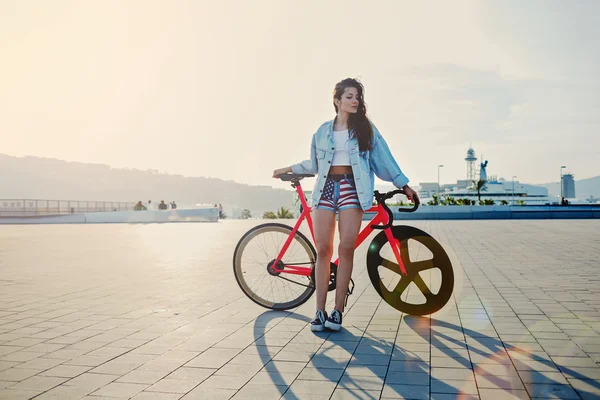 Досить молода коричнева волохата жінка стоїть зі своїм сучасним рожевим велосипедом на заході сонця, стильна дівчина-гіпстер, приваблива молода жінка виграє фіксований велосипед передач, позує на відкритому повітрі з м'яким світлом заходу сонця на фоні — стокове фото