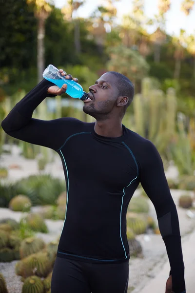 Attractive fit man in active clothes refreshing with water after run, dark-skinned healthy man with beautiful body drink water after workout outdoors, male jogger taking a break after jogging — 스톡 사진