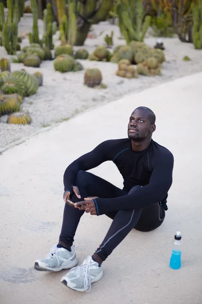 Souriant coureur à la peau foncée se reposant après l'entraînement à l'extérieur, coureur masculin en utilisant un téléphone mobile pour naviguer sur Internet pendant qu'il a la pause après l'entraînement sportif, sportif en utilisant un téléphone portable pour discuter — Photo