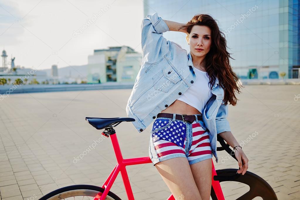 Brown haired woman posing with fixed gear bicycle outdoors, fashion girl standing with her bicycle looking to the camera
