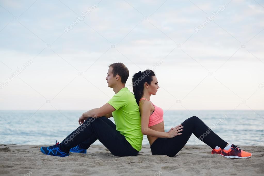 Male and female runner sitting on the sand enjoying beautiful evening, young fit couple having break after workout outdoors, beautiful couple of athletes resting after run sitting on seashore