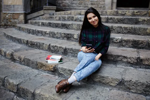 Attractive student girl resting outdoors during her class break in college, laughing female tourist girl holding mobile phone in the hand looking to the camera