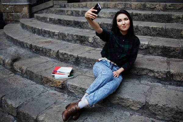 Giovane ragazza affascinante sorridente mentre prende un auto-ie all'aperto, sorridente studentessa facendo un autoritratto con smart phone seduto su gradini, bella giovane ragazza hipster fotografarsi con il telefono — Foto Stock