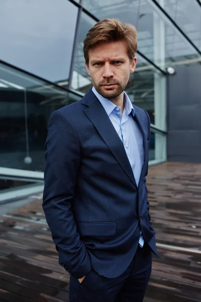 Portrait of sad businessman standing with hands in suit pockets, desperate businessman standing against skyscraper office building, portrait of defeat businessman looking to the camera — Stock Fotó