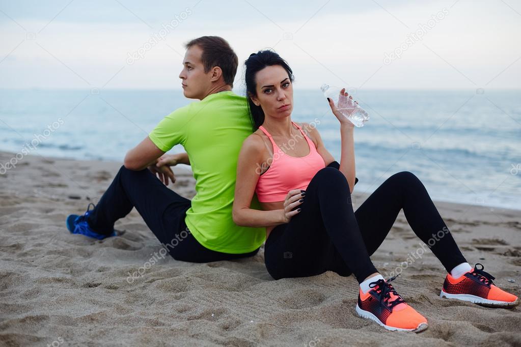 Young fit couple having break after workout outdoors, attractive brunette woman refreshing with bottle of water looking to the camera, beautiful young couple of man and woman resting after workout
