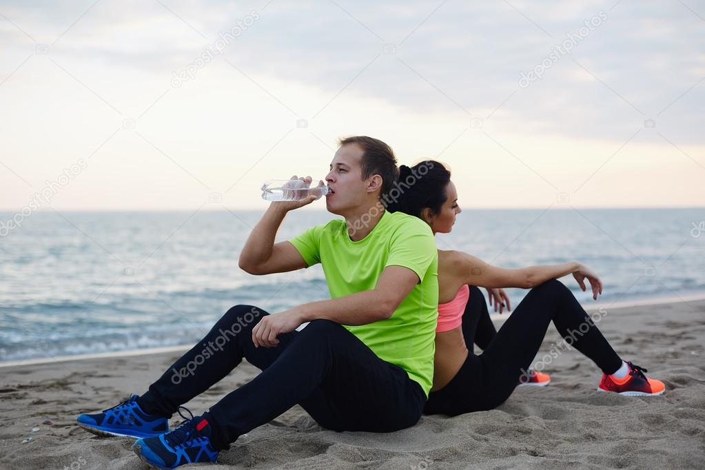 Young fit couple having break after workout outdoors, beautiful caucasian man refreshing with bottle of water exhausted after run, resting couple of two runners sitting on the beach, living healthy