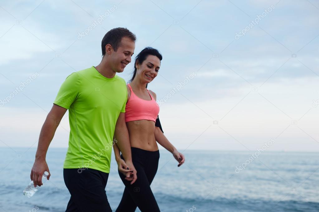 Sport couple walking along the beach resting after workout, sexy fit woman and man dressed in fluorescent-shirt taking break after run, laughing couple walk along seashore after fitness training
