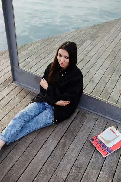 Attractive student girl seated on wooden pier having class break, female tourist woman with serious looking to the camera, stylish hipster girl with crossed arms posing outdoors