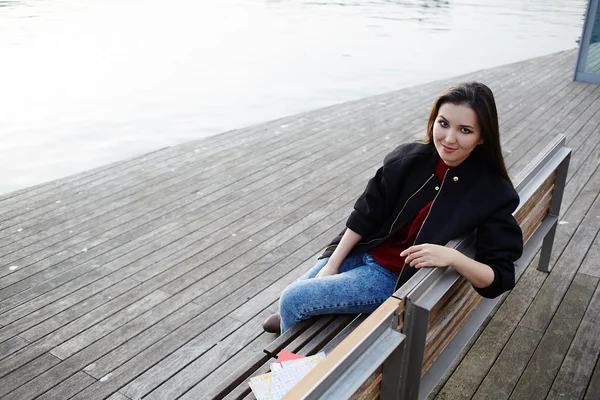 Menina estudante atraente sentada no banco de madeira com pausa de classe, mulher turista feminina com sorriso olhando para a câmera, menina hipster elegante posando ao ar livre — Fotografia de Stock