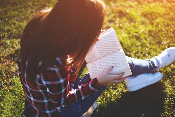 Девушка, сидящая в парке на зеленой траве и читающая книгу в красной обложке — стоковое фото