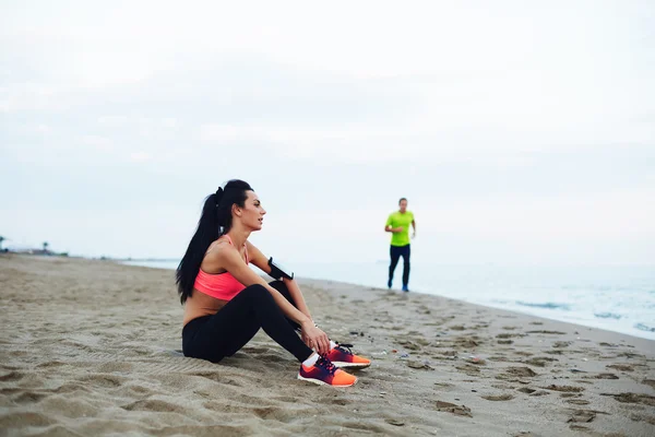 Hombre y mujer juntos practican deportes hasta que el chico corriendo chica descansando sentado en la arena —  Fotos de Stock