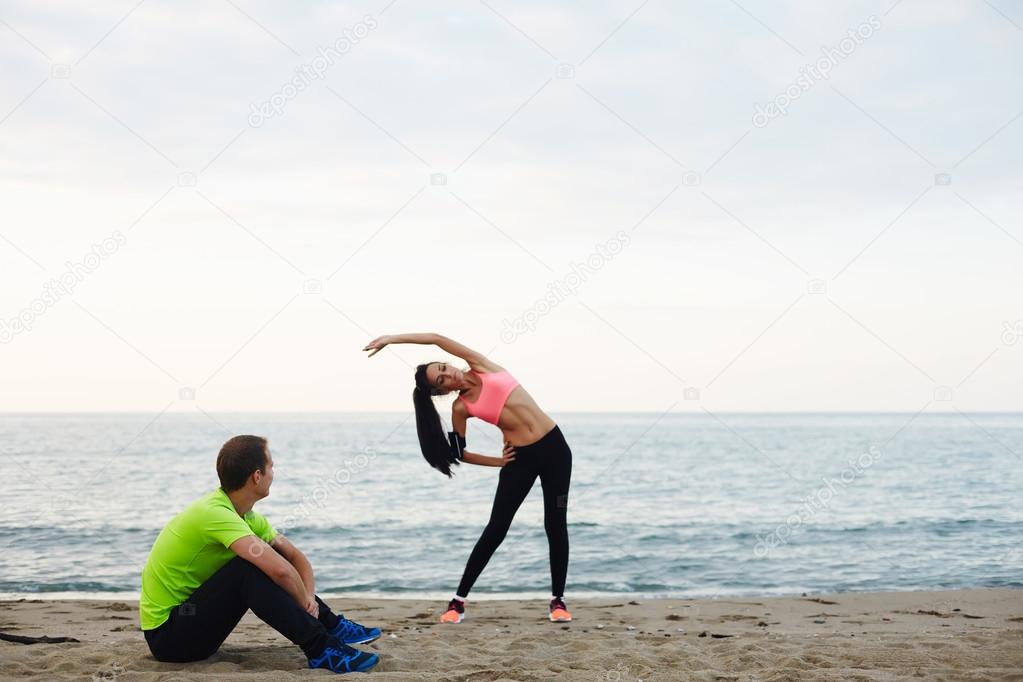 Young fitness woman doing physical stretch exercise while her boyfriend taking break