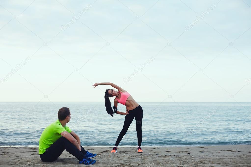The guy sitting on the sand near the water, watching as his girl doing stretching