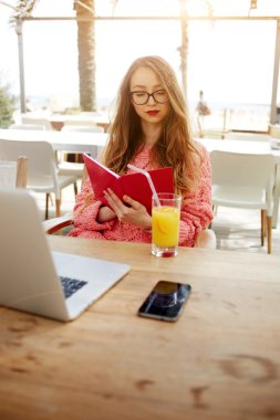 Attractive creative freelancer read notebook while sitting at wooden table with laptop laptop computer and cellphone