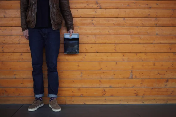 Junger Kerl mit stylischer Lederjacke steht auf dem Hintergrund einer Holzwand und hält ein digitales Tablet in der Hand — Stockfoto