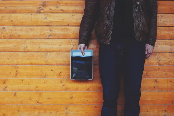 Young guy with a stylish leather jacket standing on the background of wooden wall, holding digital tablet — Zdjęcie stockowe