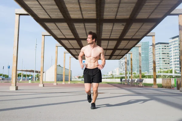 Full length portrait of handsome sportsman with good muscular build doing an intensive fitness training in urban setting, young male athlete with bottle of water in hand engaged in active sports while looking away — Stock Photo, Image