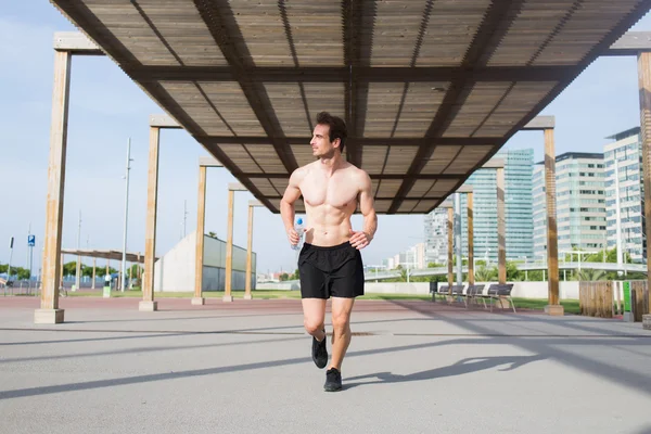 Retrato de comprimento total de belo desportista com boa construção muscular fazendo um treinamento intensivo de fitness em ambiente urbano, jovem atleta do sexo masculino com garrafa de água na mão envolvida em esportes ativos enquanto olha para longe — Fotografia de Stock