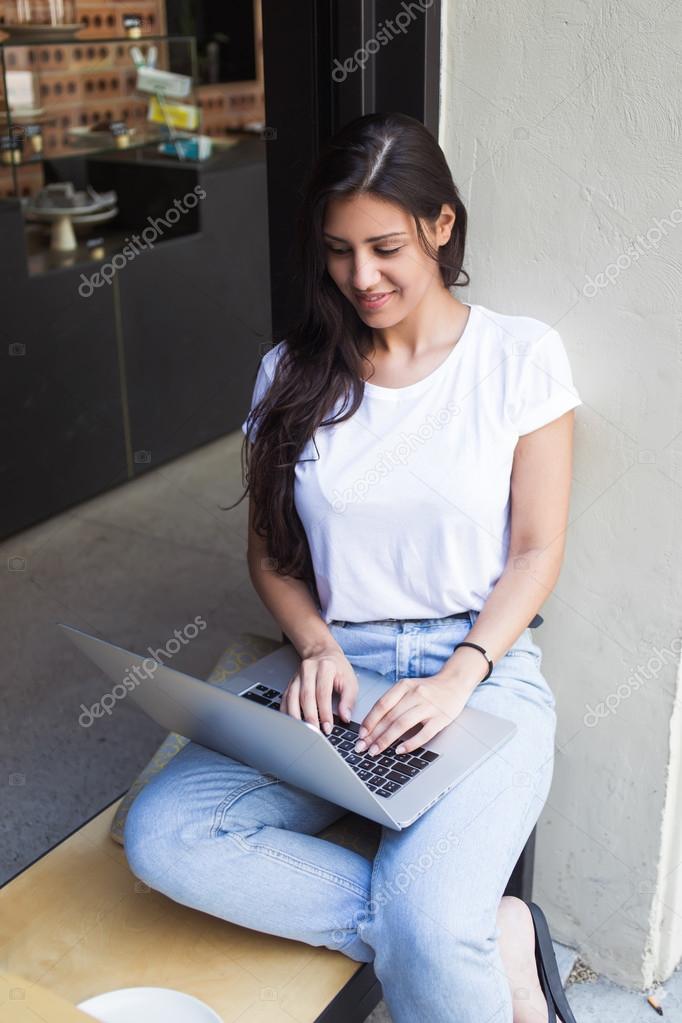 Charming female student keyboarding on her net-book while resting in coffee shop after lectures, young latin woman using laptop computer for distance work while sitting in sidewalk cafe outdoors