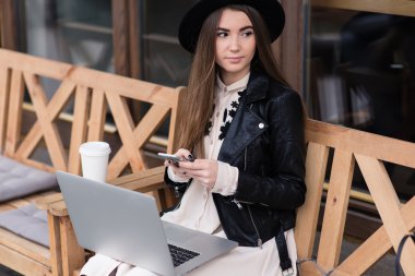 Smiling trendy woman using smart phone while sitting with laptop in sidewalk cafe