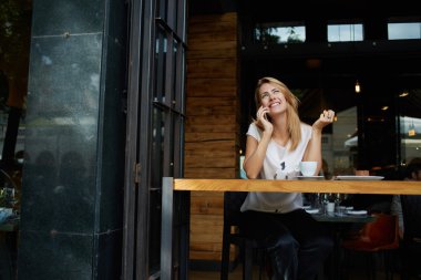 Young pretty female with good mood talking on mobile smart phone with her friend while relaxing in cafe, happy woman speaking with cell telephone while sitting in coffee shop during morning breakfast