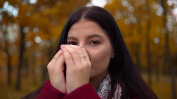 Жінка дивиться на камеру, п'є гарячий кавовий чай на відкритому повітрі, осінь гуляє в парку — стокове відео