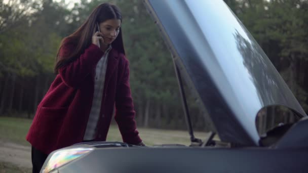 Woman talking by phone, looking at broken car motor, calls to evacuation service — Stock Video