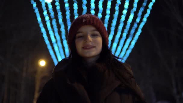 笑顔の女性はカメラを見て、通り、冬の夜、街路灯を歩く — ストック動画