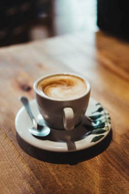 Tahta bir masanın üzerine yeşil yapraklar çizilmiş beyaz bir bardaktaki kahvenin ön görüntüsü. Kahvaltı konsepti.