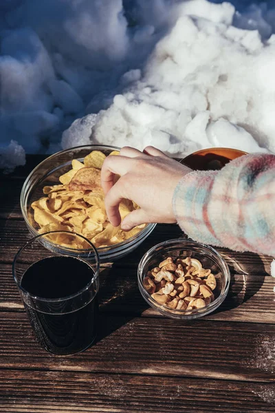 A woman\'s hand grabs a potato chip from a snack table on a rustic board in the snow. Winter concept. Food concept.