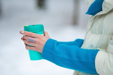Bir kış günü, arka planda beyaz bir karla yeşil bir termal bardak tutan bir kadının elleri..