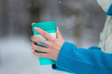 Bir kış günü, arka planda beyaz bir karla yeşil bir termal bardak tutan bir kadının elleri..