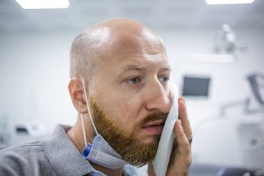 Mutsuz ve korkmuş beyaz bir erkek diş hekiminde diş ağrısı yüzünden yanağında soğutma maddesi tutuyor ve muayene olmayı bekliyor.. 