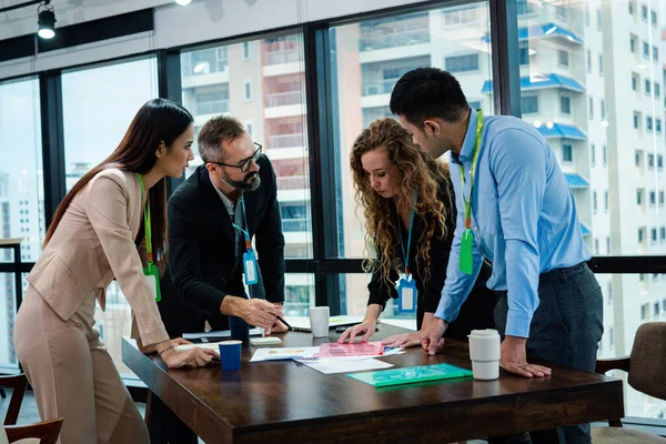 Business team are in the brainstorming stage to plan about the marketing of the project. They are standing while they discuss the project. There are lots of the document, note book on the table.